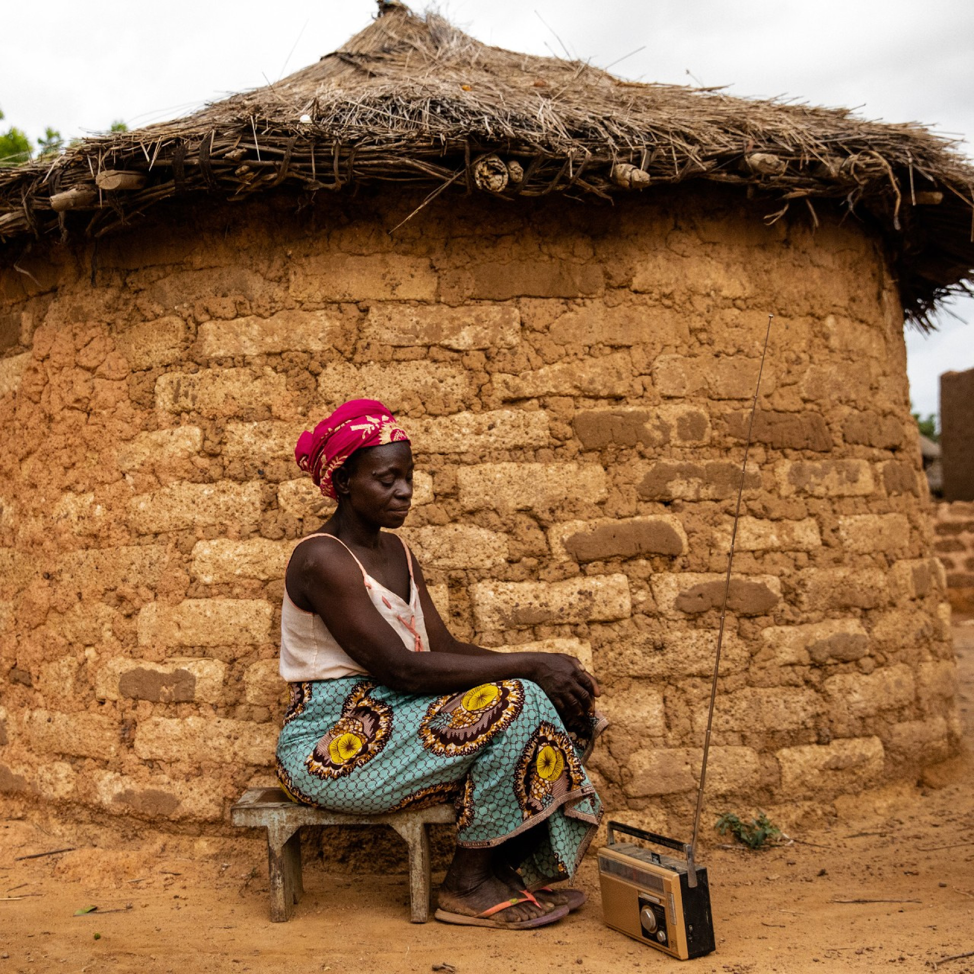 A woman sitting by a hut with a radio resembling the far reach of DMI radio campaigns